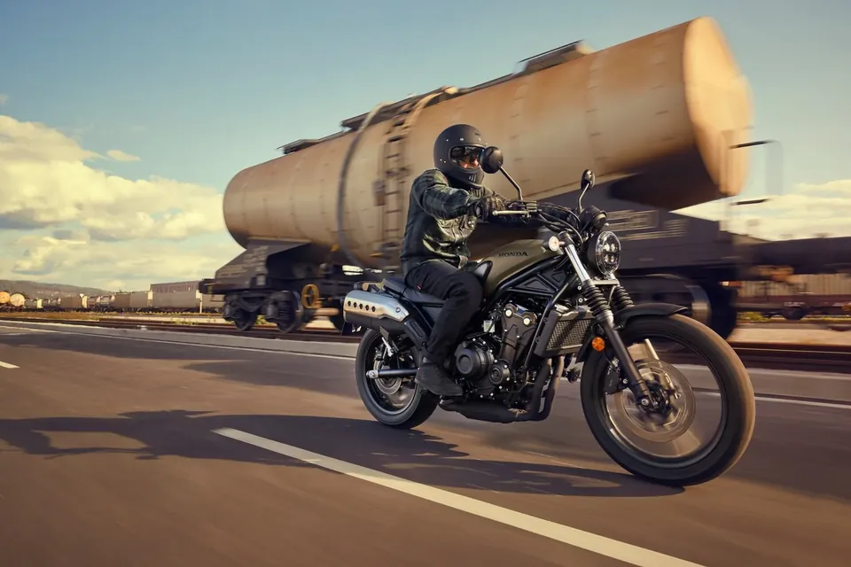 Honda CL500 avec pilote sur une route de campagne devant un camion-citerne